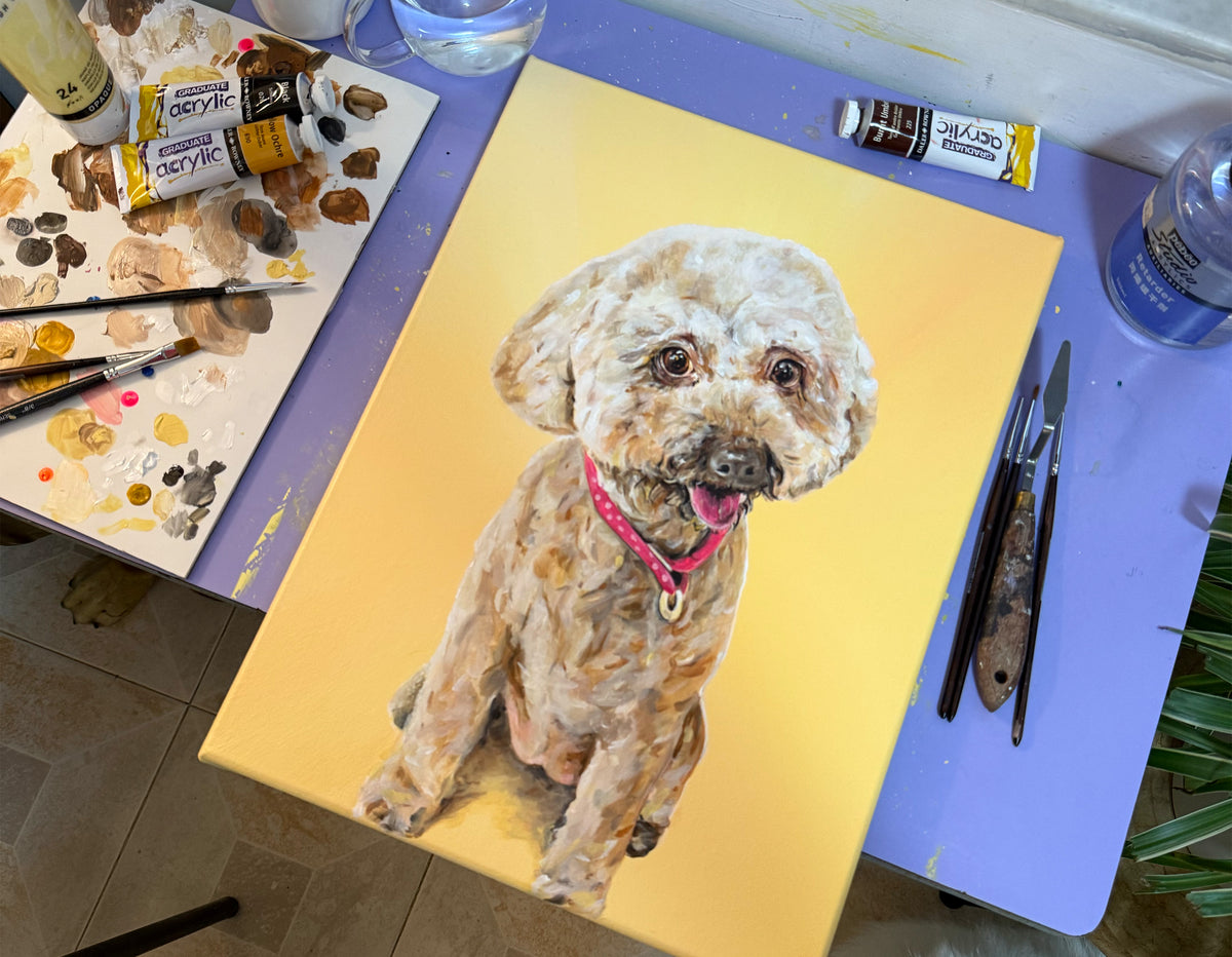 A custom pet portrait canvas showing a painted fluffy dog with a pink collar on a yellow background, displayed on a purple artist table with paint tubes and brushes.